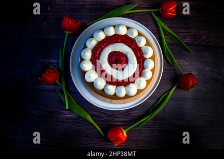 Honigschwammkuchen mit einer Dekoration aus weißer Luftcreme und Schichten aus roten Himbeeren steht auf einer weißen Platte auf einem dunklen Holzhintergrund, Tulpen liegen Stockfoto