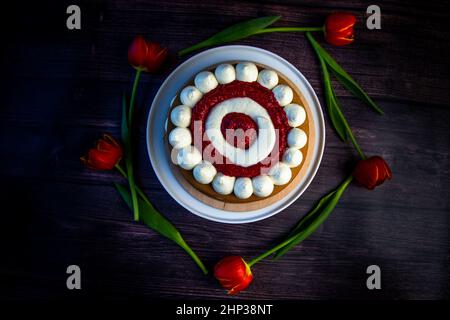 Honigschwammkuchen mit einer Dekoration aus weißer Luftcreme und Schichten aus roten Himbeeren steht auf einer weißen Platte auf einem dunklen Holzhintergrund, Tulpen liegen Stockfoto