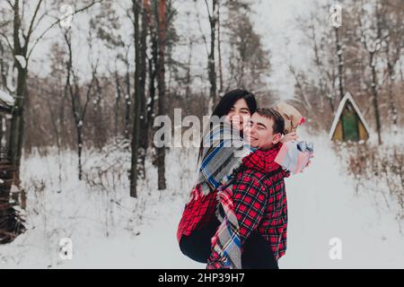 Freund gibt eine Geschenkbox Geschenk mit Band im Freien aufgrund der heiligen Valentinstag zu ihrer Freundin. Verschneite Wälder, Bäume mit Schnee. Zwei sind gekleidet i Stockfoto