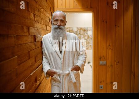 Lächelnder bärtiger Mann im Bademantel, der sich im Saunabereich aus Holz im Spa-Resort entspannt und die Kamera anschaut Stockfoto