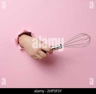 Weibliche Hand hält einen Metallbesen, um Produkte auf einem rosa Hintergrund zu schlagen. Ein Teil des Körpers ragt aus einem zerrissenen Loch heraus Stockfoto
