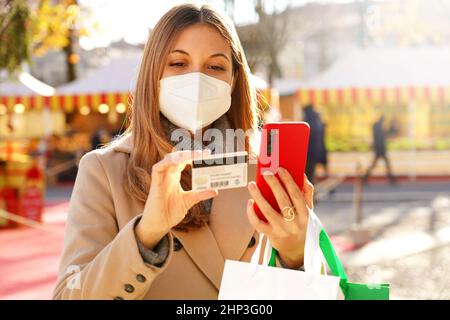 Attraktive Shopper Frau trägt medizinische Gesichtsmaske beobachten ihre Kreditkartennummer für Online-Shopping auf Handy mit verschwommenem weihnachten Backgrou Stockfoto