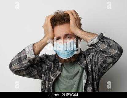 Kranker junger Mann in medizinischer Gesichtsmaske fühlt sich Migräne, die den Kopf hält. Schöner Kerl fühlen Schmerzen oder Kopfschmerzen. Erschöpfter junger Mann, der versucht, sich zu konzentrieren und seinen Kopf massiert. Gesundheitskonzept. Covid 19-Konzept. Stockfoto