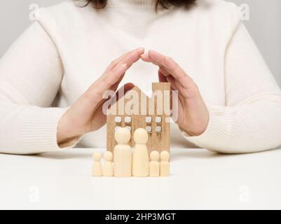 Ein Holzhaus und Figuren einer Familie, zwei weibliche Palmen schützen Objekte von oben. Immobilienversicherungskonzept, glückliche Familie Stockfoto