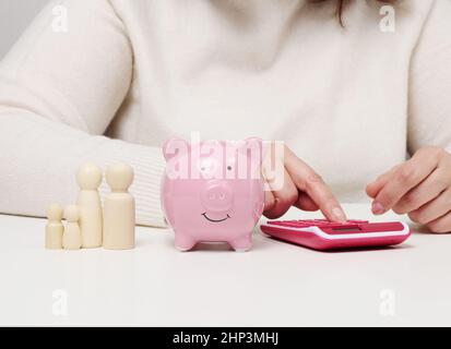 Pinkfarbenes Sparschwein aus Keramik, Holzfiguren aus der Familie. Frau sitzt am Tisch und zählt auf Rechner, Budget-Planung Konzept Stockfoto