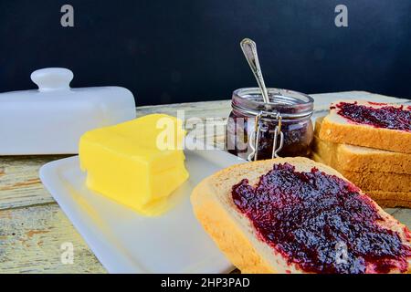 Marmelade, Butter in Butterform und Marmelade auf Toast verteilen. Gesund und Diät-Konzept. Ländlicher weißer Holzhintergrund. Stockfoto