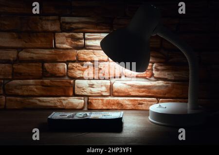 Ein Stapel von hundert-Dollar-Scheinen auf einem Holztisch wird vom Licht einer Lampe vor dem Hintergrund einer Ziegelmauer beleuchtet. Stockfoto