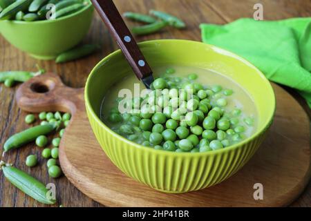 Frische grüne Erbsensuppe mit Erbsensamen und Erbsenschoten herum. Selektiver Fokus, Fokus auf die Erbse in der Mitte der Suppe. Stockfoto