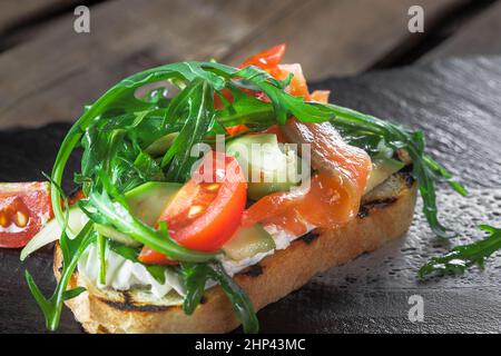 Bruschetta mit Lachs, Avacado, Tomate und Rucola, mit cremiger Sauce, liegt auf einem Steinständer, Nahaufnahme Stockfoto