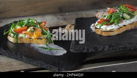 Zwei Bruschettas mit Lachs und Roastbeef, mit Sahnesauce, Tomaten und Rucola, liegen auf Steinstützen auf einer Holzoberfläche Stockfoto