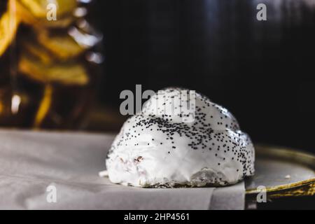 Bun in Form einer Rutsche, verziert mit weißer Buttercreme und Mohnsamen, steht auf Pergament auf einem Eisenblech, dunkler Hintergrund Stockfoto