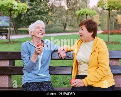 Zwei reife Frauen essen Äpfel auf der Bank nach dem Sport-Übungen im Park. Gesundes Lifestyle-Konzept Stockfoto