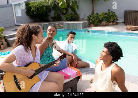 Multirassische Freunde hören jungen Frauen am Pool akustische Gitarre spielen an sonnigen Tag Stockfoto