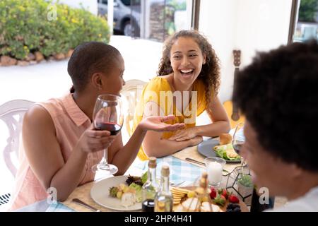 Fröhliche junge weibliche multirassische Freunde lachen zusammen, während sie am Esstisch sitzen Stockfoto