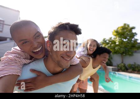 Junge Birazialmänner, die fröhlichen Frauen am Pool an sonnigen Tagen Huckepack-Fahrten geben Stockfoto