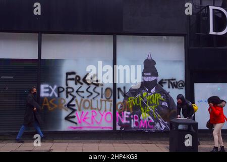 Manchester, Großbritannien, 18th. Februar 2022. Ein humorvolles Wandgemälde mit dem britischen Premierminister Boris Johnson im alten Debenhams-Laden im Zentrum von Manchester, Großbritannien, wurde bisher zweimal mit Graffiti verwüstet. Produziert von 'Foka Wolf', einem Straßenkünstler, hat das Wandbild Boris Johnsons Kopf, trägt einen Partyhut, auf einem Bild eines bewaffneten Polizisten. Während der Covid-19-Sperre gibt es eine polizeiliche Untersuchung von Parteien in der Downing Street 10. Der Künstler hat erwähnt, was er als die "doppelten Standards" der Regierung ansieht. Die Menschen gehen an dem Wandbild vorbei. Quelle: Terry Waller/Alamy Live News Stockfoto