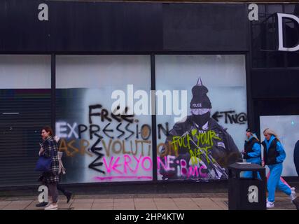 Manchester, Großbritannien, 18th. Februar 2022. Ein humorvolles Wandgemälde mit dem britischen Premierminister Boris Johnson im alten Debenhams-Laden im Zentrum von Manchester, Großbritannien, wurde bisher zweimal mit Graffiti verwüstet. Produziert von 'Foka Wolf', einem Straßenkünstler, hat das Wandbild Boris Johnsons Kopf, trägt einen Partyhut, auf einem Bild eines bewaffneten Polizisten. Während der Covid-19-Sperre gibt es eine polizeiliche Untersuchung von Parteien in der Downing Street 10. Der Künstler hat erwähnt, was er als die "doppelten Standards" der Regierung ansieht. Die Menschen gehen an dem Wandbild vorbei. Quelle: Terry Waller/Alamy Live News Stockfoto