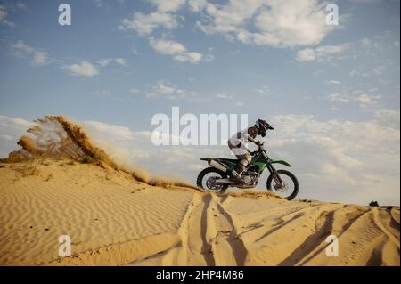 Professioneller Motocross-Fahrer auf Helm und Schutzanzug, der den Sandberg hinunterrutscht. Speedmoto-Rennen in der Wüste Stockfoto