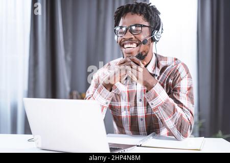 Glücklicher Freiberufler oder afroamerikanischer Student, der zu Hause mit einem Laptop und Kopfhörern für einen Videoanruf in einer Gruppe arbeitet, der entfernt arbeitet. Junge Studentin auf entfernter Neigung hat einen Video-Gruppenanruf. Stockfoto