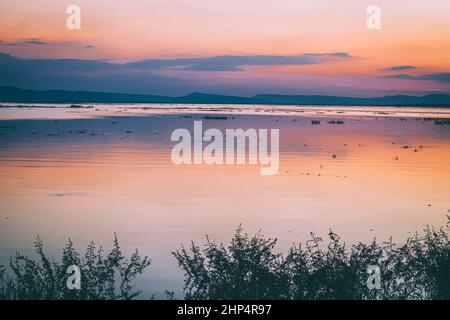 Lange Belichtung Vanille bunten Himmel und See Reflexion mit Bergen im Hintergrund. Verschwommenes wildes Gras weht im Wind im Vordergrund Stockfoto