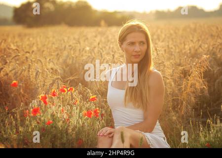 Junge Frau im weißen Kleid, sitzen im Weizenfeld, roter Mohn in der Nähe von Sonnenuntergang leuchtet. Stockfoto