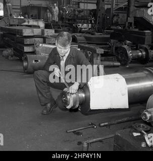 1950s, historisch, in einer Industriewerkstatt in einer Stahlfabrik, ein junger Ingenieurslehrling in einem Hemd und Krawatte und Arbeitskleidung, der sich an einem großen industriellen Stahlteil hockend, mit einer Handfeile, um seine Vorderkante zu glätten, Port Talbot, Wales, Großbritannien. Neben ihm einige technische Zeichnungen. Stockfoto