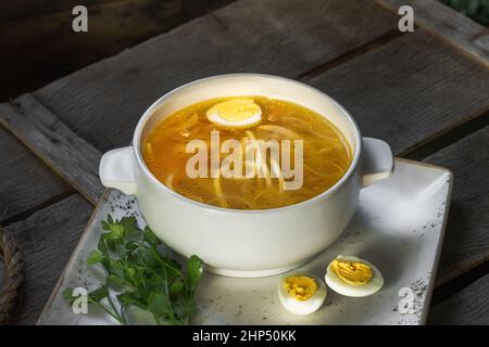 Suppe mit Nudeln und Hühnerfleisch in einem weißen Porzellanteller steht auf einem Keramiktablett, daneben ein geschnittenes gekochtes Hühnerei und Petersilie, Draufsicht Stockfoto