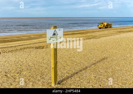 Vorsicht vor dem Cliffing-Schild am Snettisham Strand in Norfolk mit einem Volvo A30G-Muldenkipper im Hintergrund, der am Ufer des Wash Strandsanierungsarbeiten durchführt. Stockfoto