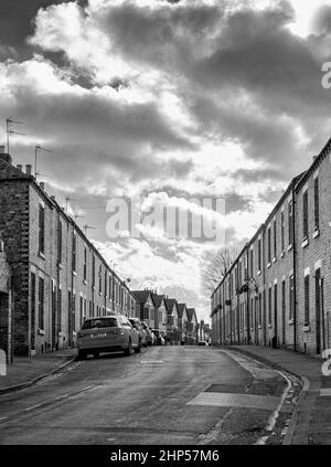Eine Straße mit gepflegten Reihenhäusern liegt auf einem nach oben abfallenden Anstieg. Autos werden auf einer Seite geparkt und ein wolkiger Himmel ist oben Stockfoto