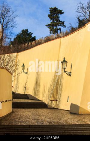 Ein gepflasterter Durchgang mit der gelben Pfeilermauer der Prager Burg und einer traditionellen verzierten Straßenlaterne, an einem sonnigen Tag mit Schatten, Prag, Tschechien Stockfoto