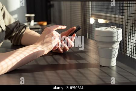 Weibliche Hände in Nahaufnahme halten das Telefon. Frau, die am Cafe-Schreibtisch mit Öko-Tasse und berührender Telefonanzeige sitzt. Verwendung von Smartphones im Common Life-Konzept. Hochwertige Fotos Stockfoto