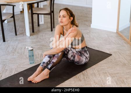 Junge weiße Frau, die auf einer Yogamatte sitzt, während sie im Zimmer arbeitet Stockfoto