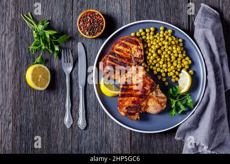 Saftig gegrillte Schweinefleisch-T-Bone-Steaks mit gekochten grünen Erbsen und Zitrone auf Teller auf einem Holztisch mit Besteck, horizontale Ansicht von oben, flach legen Stockfoto