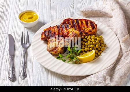 Gegrillte Schweinefleisch-T-Bone-Steaks mit gekochten grünen Erbsen und Zitrone auf einem weißen Teller auf einem Holztisch mit Senf Stockfoto