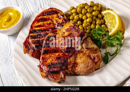 Gegrillte Schweinefleisch-T-Bone-Steaks mit gekochten grünen Erbsen und Zitrone auf einem weißen Teller auf einem Holztisch, horizontale Ansicht von oben, Nahaufnahme Stockfoto