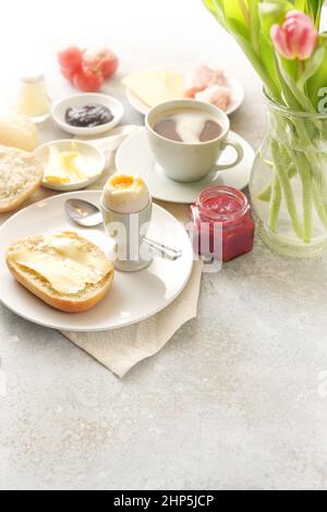 Frühstück mit Brötchen, Ei, Marmelade und einer Tasse Kaffee, heller Hintergrund mit einer Blumenvase, mit Liebe am Muttertag oder Geburtstag serviert, Copy space, vert Stockfoto