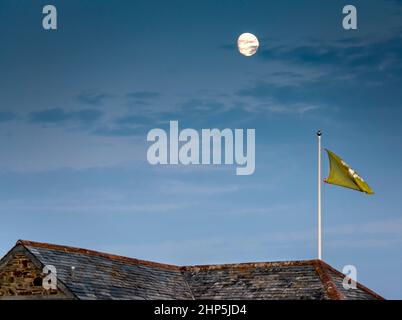 Eine Flagge des National Trust fliegt hoch auf einem kornischen Dach, und der silberne Mond leuchtet darüber hinaus vor einem dunklen Himmel. Stockfoto
