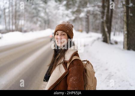 Glückliche asiatische weibliche Reisende, die entlang der Autobahn entlang der Autobahn entlang schneereichen Winterwald trampeln Stockfoto