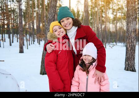 Entzückte liebevolle Mutter - schöne Frau umarmte sanft ihre entzückenden Kinder, Junge und Mädchen, lächelnd mit fröhlichem toothy Lächeln, während sie auf einem sno gehen Stockfoto