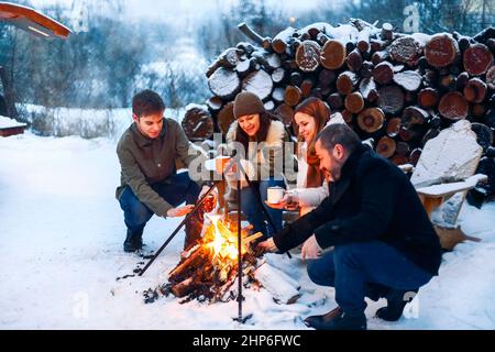 Eine Gruppe von Freunden, die sich am Lagerfeuer im Hinterhof treffen, Tee trinken und sich die Hände wärmen. Zwei glückliche Paare, die sich entspannen und die Wintersaison genießen, während sie sitzen Stockfoto