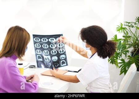 afroamerikanische Ärztin mit Gesichtsmaske, die einer Patientin während eines medizinischen Termins in einer Klinik oder einem Krankenhaus eine CD mit Röntgenbild übergibt, selecti Stockfoto