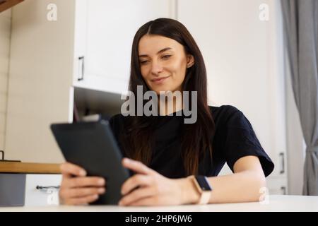 Fröhliche weiße Frau, die auf einem Laptop in der heimischen Küche im Internet surfen kann. Glückliche Brünette weiblich mit langen dunklen Haaren mit modernen Gerät für Online-Co Stockfoto
