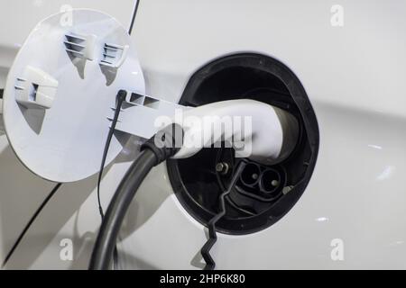 Nahaufnahme des elektrischen Ladegerätes, der mit einem Stecker zum Laden der Batterie am weißen EV-Auto verbunden ist Stockfoto