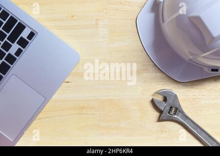 Laptop mit Nahaufnahme, weißer Sicherheitshelm und Schraubenschlüssel auf Holztisch mit Platz für Kopien in der Mitte auf der Draufsicht Stockfoto