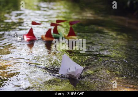 Schiffe aus Küchenschwämmen auf der Suche nach einem Weißpapierschiff. Segelbootregatta für Kinder. Selbstgemachte Kinderboote, die auf einem kleinen Fluss oder einer Straße segeln Stockfoto