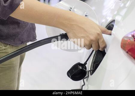 Nahaufnahme eines Mannes, der einen wiederaufladbaren Gerätestecker für die Ladebatterie an der Buchse eines weißen Elektroautos hält, Stockfoto