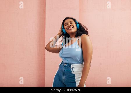 Fröhliche Frau mit schwarzen lockigen Haaren, die blaue Kopfhörer an der rosa Wand trägt Stockfoto
