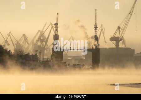 Kränen der Werft in frostigen Wintertag, Dampf über dem Fluss, glatte Oberfläche des Flusses bei Sonnenuntergang. Stockfoto