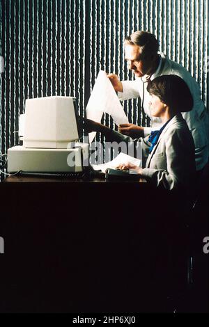 Eine weibliche Bibliothekarin, die an einem Computer sitzt, der mit PDQ arbeitet, während ein männlicher Arzt einen Ausdruck untersucht. 1989 Stockfoto