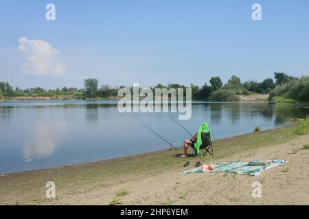 Fischer sitzt am Ufer eines Flusses und fängt Fische mit zwei Angelruten. Rückansicht, selektiver Fokus. Stockfoto